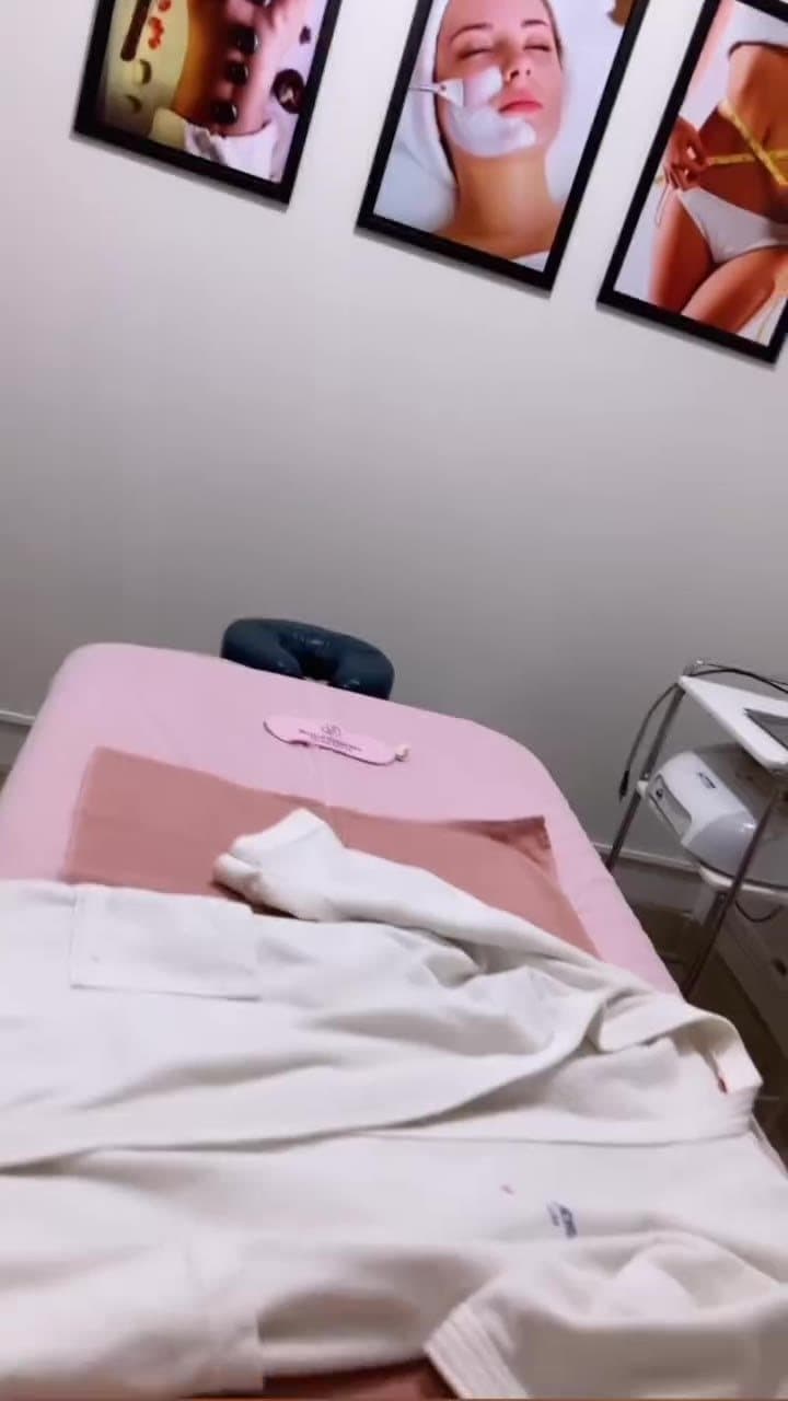 Sala de estética com maca forrada de rosa, roupão branco e quadros na parede